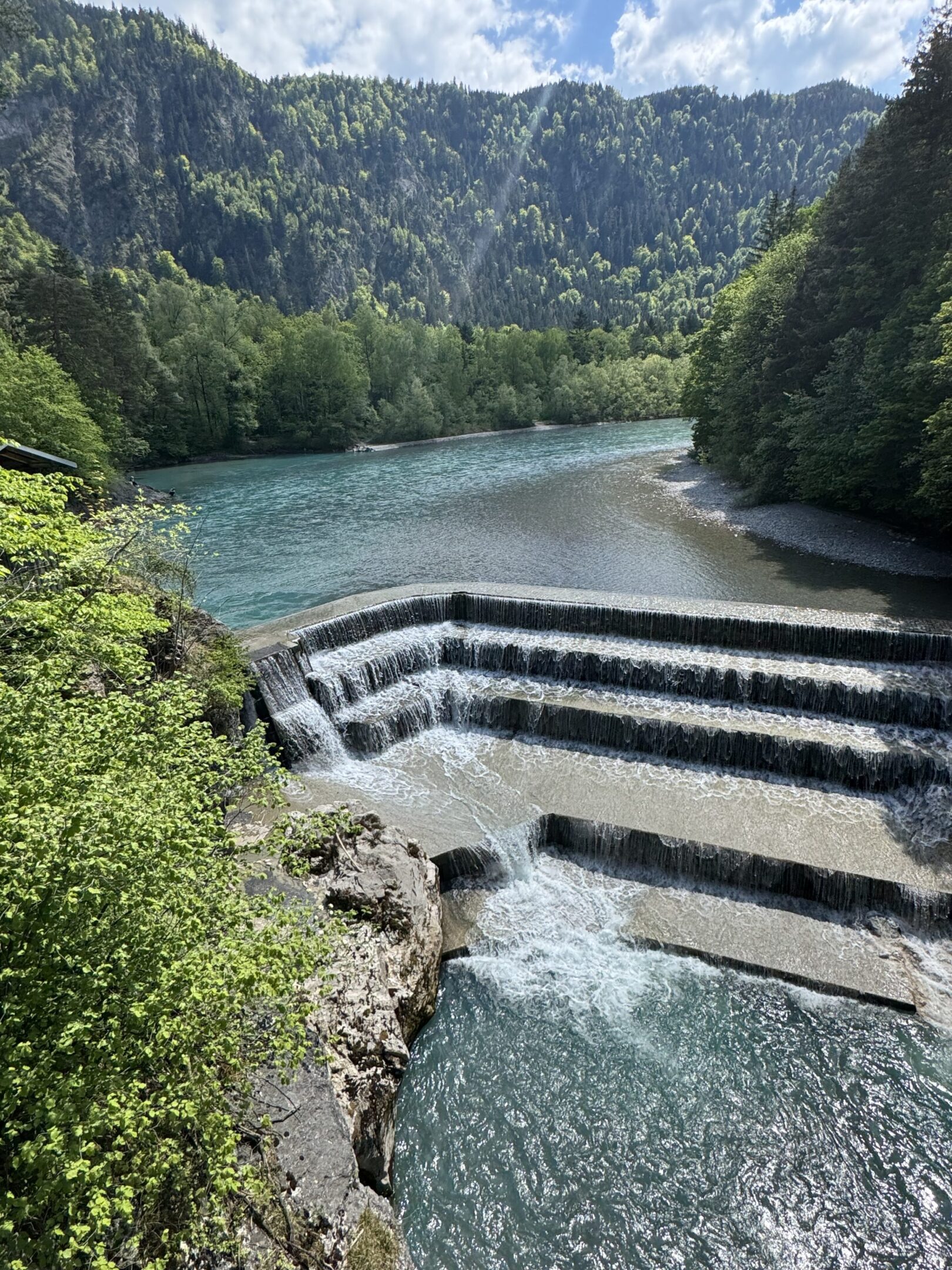 A stepped waterfall flows into a clear river surrounded by green trees and mountains.