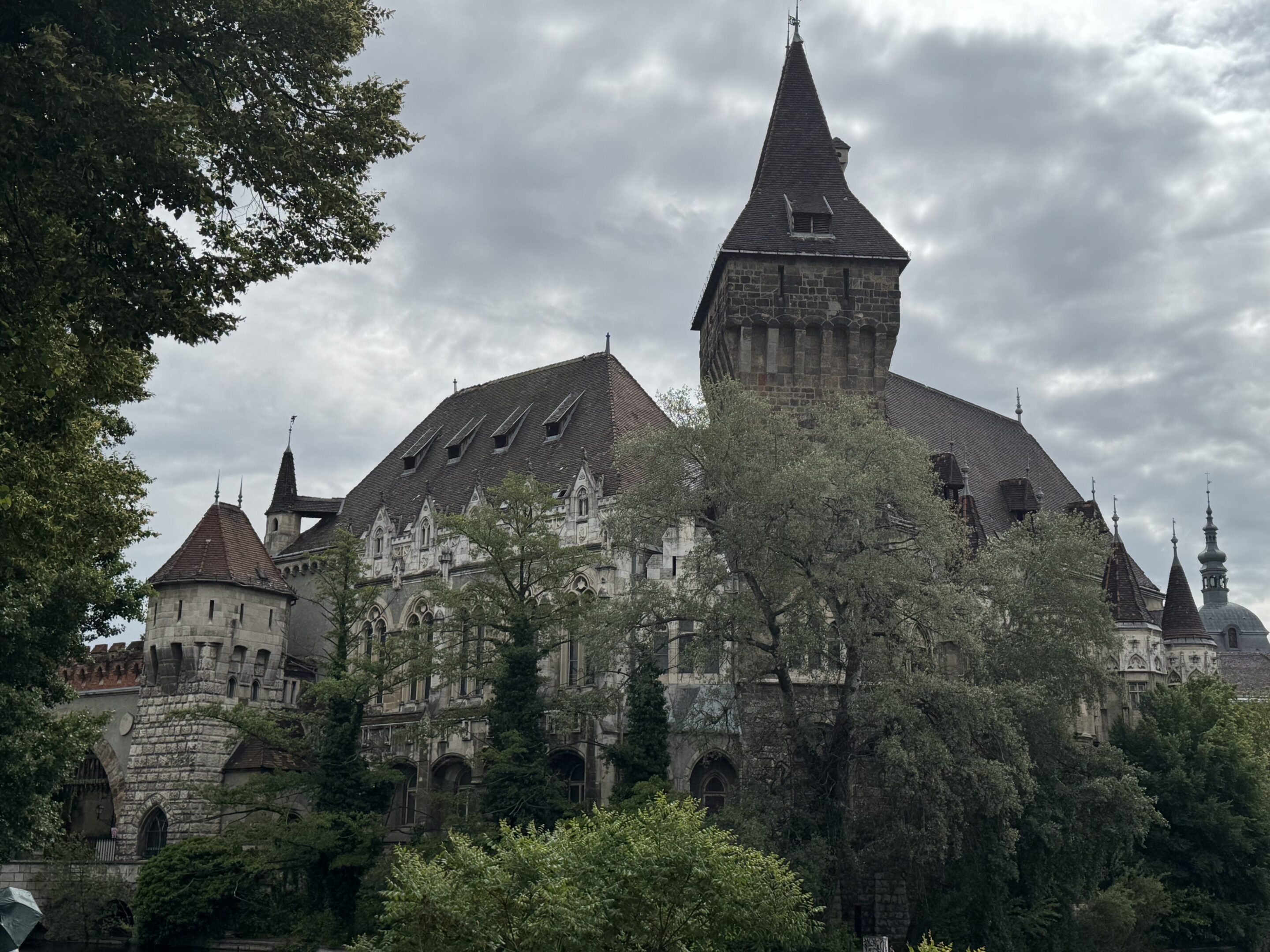 Historic castle with tall spires surrounded by trees under a cloudy sky.