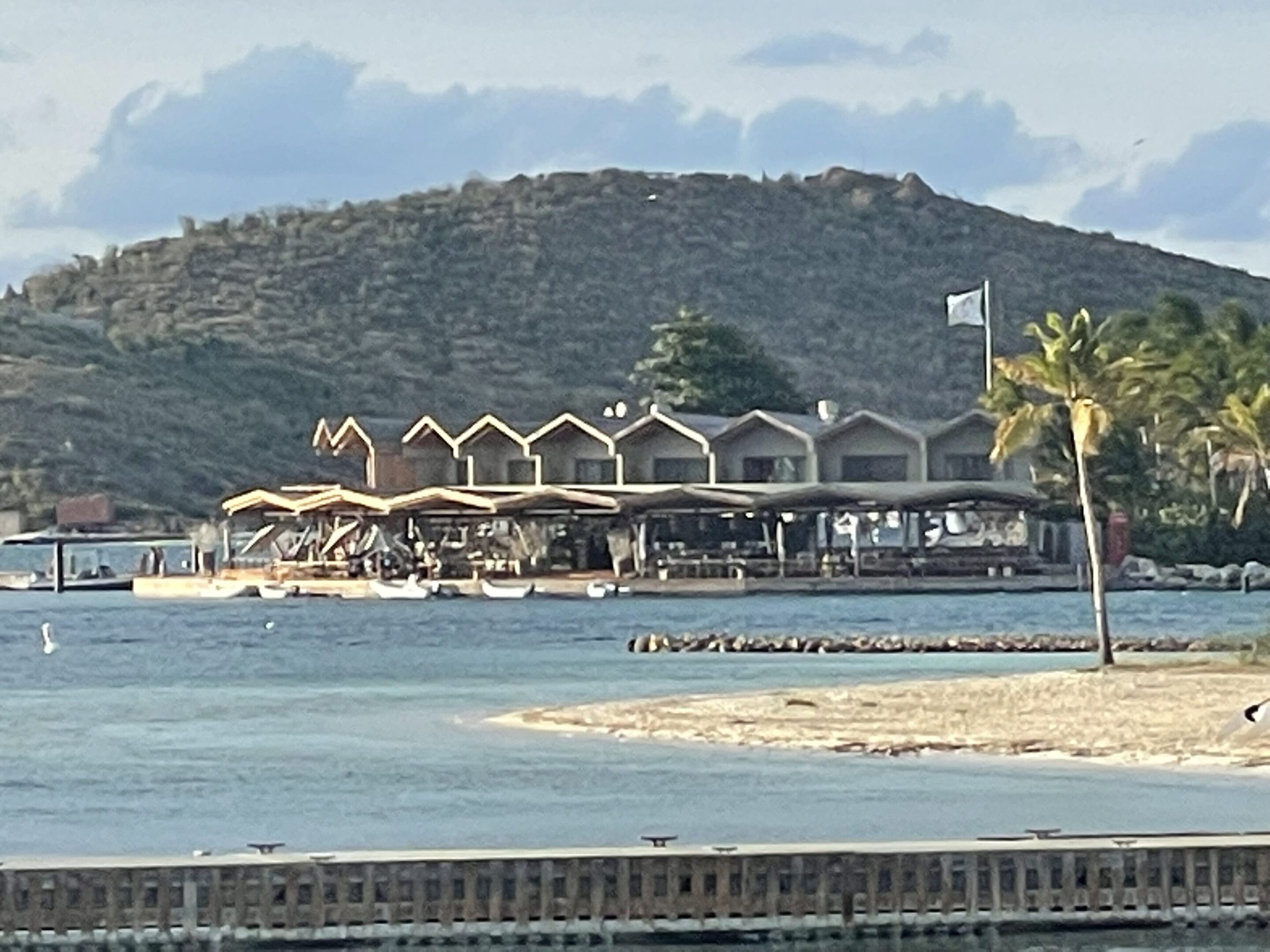 Beachside restaurant with umbrellas and seating near calm waters and hills.