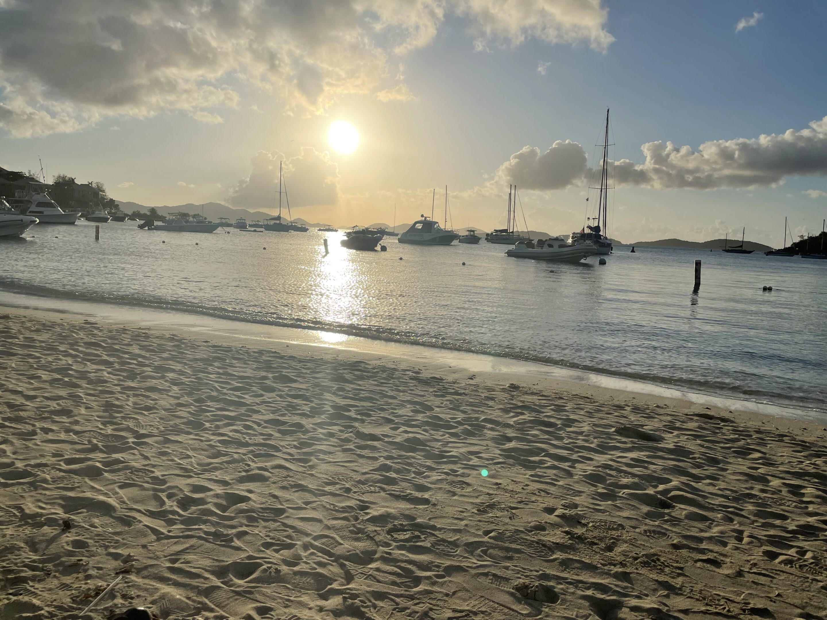Sunset over a calm beach with sailboats on the water.
